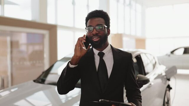 Car dealer holding clipboard in automobile showroom. Smiling salesman using smartphone while having business call at car showroom. Professional confident sales person working in modern car dealership.