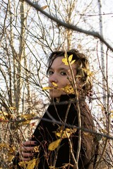 Thoughtful young woman standing behind tree branches in autumn forest - 791434887