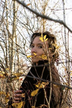 Thoughtful young woman standing behind tree branches in autumn forest