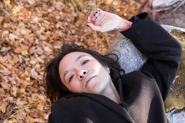 Thoughtful young woman lying on autumn leaves and looking at camera - 791435282