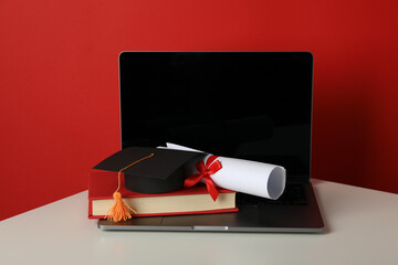 Graduate hat with diploma, book and laptop on red background.