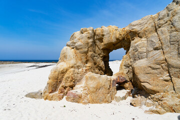 Plage de sable blanc bordée par une arche naturelle creusée dans la roche, baignée par des eaux turquoises, sous un ciel bleu en Finistère Nord, Bretagne. - obrazy, fototapety, plakaty