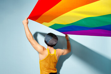 Back rear view young happy gay Latin man wear mesh tank top hat clothes hold in hand striped rainbow flag isolated on plain blue cyan background studio portrait Pride day June month love LGBT concept