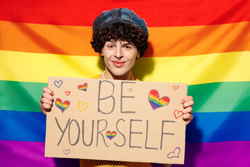 Young happy gay Latin man wears mesh tank top hat clothes hold card sign with be yourself title...