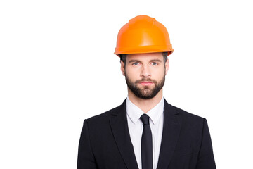 Portrait of concentrated, thoughtful architector with stubble in orange safety helmet, hardhat and black tux with tie, looking at camera, isolated on grey background