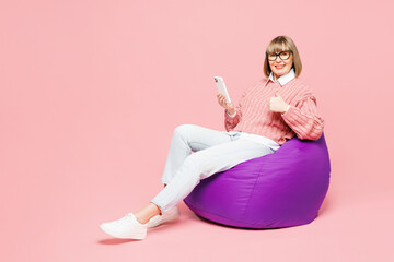 Full body elderly woman 50s years old wear sweater shirt casual clothes glasses sit in bag chair hold use mobile cell phone show thumb up isolated on plain pink background studio. Lifestyle concept.