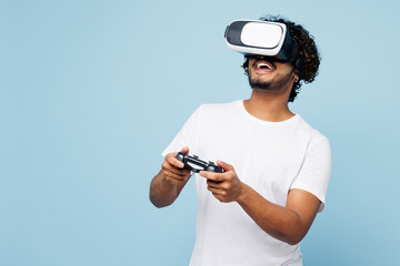 Young happy Indian man he wear white t-shirt casual clothes watching in vr headset pc gadget play pc game with joystick console isolated on plain pastel light blue cyan background. Lifestyle concept.