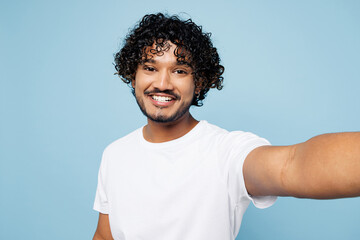 Close up young happy Indian man he wear white t-shirt casual clothes doing selfie shot pov on mobile cell phone isolated on plain pastel light blue cyan background studio portrait. Lifestyle concept.