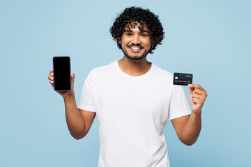 Young Indian man wear white t-shirt casual clothes using blank screen mobile cell phone credit bank card shopping online order delivery book tour isolated on plain blue background. Lifestyle concept.