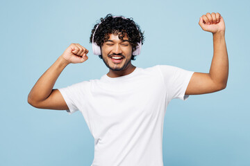 Young happy Indian man he wears white t-shirt casual clothes listen to music in headphones raise up hands dance isolated on plain pastel light blue cyan background studio portrait. Lifestyle concept.