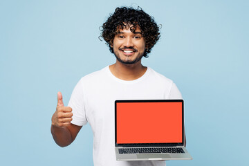 Young happy IT Indian man he wear white t-shirt casual clothes hold use work on laptop pc computer with blank screen workspace area show thumb up isolated on plain pastel light blue cyan background.