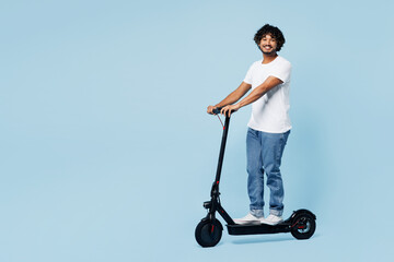 Full body young happy smiling Indian man he wear white t-shirt casual clothes ride electric scooter look camera isolated on plain pastel light blue cyan background studio portrait. Lifestyle concept.