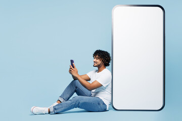 Full body fun young Indian man wear white t-shirt casual clothes sit near big huge blank screen mobile cell phone with area use smartphone isolated on plain pastel blue background. Lifestyle concept.