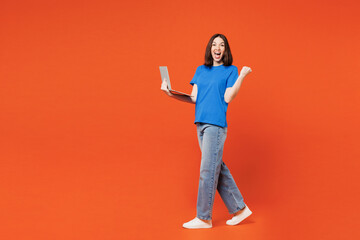 Full body young smiling fun happy IT woman she wear blue t-shirt casual clothes hold use work on laptop pc computer walk go isolated on plain red orange background studio portrait. Lifestyle concept.
