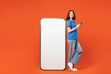 Full body side view young woman wear blue t-shirt casual clothes big huge blank screen mobile cell phone with workspace copy space mockup area using smartphone isolated on plain red orange background.