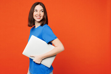 Side view young smiling happy IT woman she wear blue t-shirt casual clothes hold closed laptop pc computer look aside on area isolated on plain red orange background studio portrait Lifestyle concept