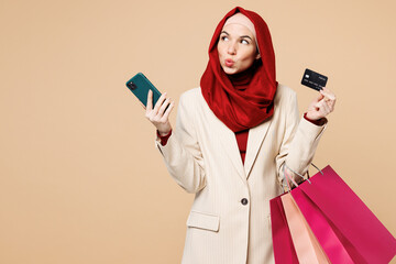 Young Arabian Asian Muslim woman wear red abaya hijab suit clothes shopping hold package bag mobile cell phone credit card isolated on plain beige background. Black Friday sale buy, UAE Islam concept.