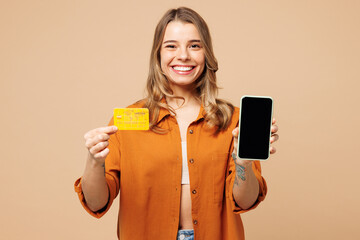 Young woman wear orange shirt casual clothes use blank screen mobile cell phone hold credit bank card shopping online order delivery booking tour isolated on plain beige background. Lifestyle concept.