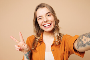 Close up young fun woman she wear orange shirt casual clothes doing selfie shot pov on mobile cell phone show v-sign isolated on plain pastel light beige background studio portrait. Lifestyle concept.