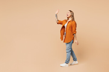 Full body side profile view young happy woman she wear orange shirt casual clothes walking going waving hand isolated on plain pastel light beige color background studio portrait. Lifestyle concept. - 791453891
