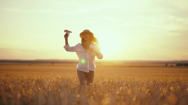 Carefree woman running on wheat field throwing paper plane in sky keeping straw hat on head at sunset, back view. Freedom, farm lifestyle, resting in countryside village, golden hours concept.