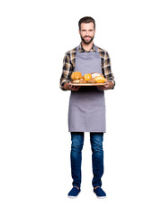 Full size body portrait of joyful cheerful baker in jeans, shoes, shirt, apron with stubble having, showing tray with bakery products, looking at camera, isolated on grey background