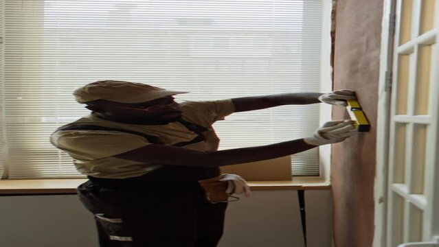 Vertical side footage of African American repairman in cap working in renovated apartment and using spirit level while making marks on wall before hanging furnitureVertical side footage of African Ame