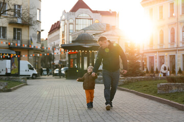 A happy three year old son walks hand in hand with his dad in the city on a sunny spring day....