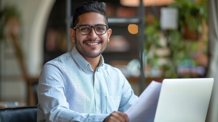 A smiling Latin American or Indian Man, businessman, Entrepreneur, Analyst, accountant, wearing a shirt, holding documents in his hands, working on a laptop, looking at a camera in a modern office.