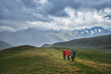 Three hikers with backpacks trek along a grassy ridge against a backdrop of dramatic mountain peaks and cloudy skies