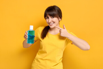 Young woman with mouthwash pointing at her healthy teeth on yellow background