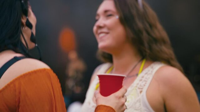 Two female friends wearing glitter holding drinks having fun dancing at outdoor summer music festival - shot in slow motion 