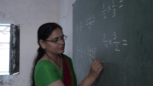 Indian female math teacher writes with chalk on the blackboard, teaching sums to elementary students. Rural india education.