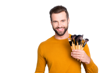 Portrait of handsome cheerful visagist with stubble in sweater having verity of brushes in arm, looking at camera, standing over grey background