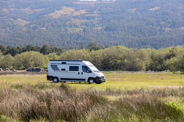 A modern white camper van is parked on a grassy field with hills and wind turbines in the...