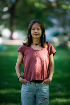 A young woman in a T-shirt and ripped jeans poses for a waist-up portrait, standing confidently in a park.