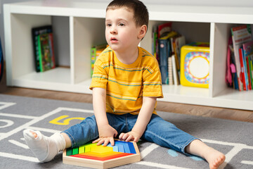 Toddler boy with a bandage or cast on his leg plays with wooden blocks. Fracture of a foot and...