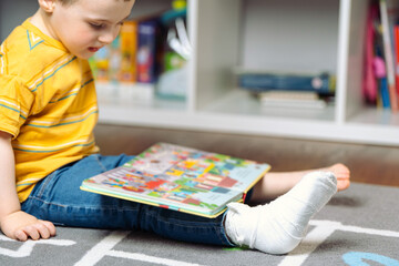 Toddler boy with a bandage or cast on his leg plays with colourful book. Fracture of a foot and...