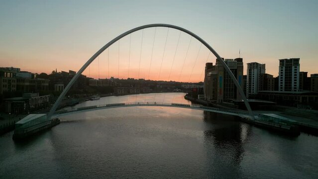 Drone slowly rises over the River Tyne as the sun rises behind the Millennium Bridge and Baltic Centre. Newcastle, Gateshead. Silhouettes cross the bridge 