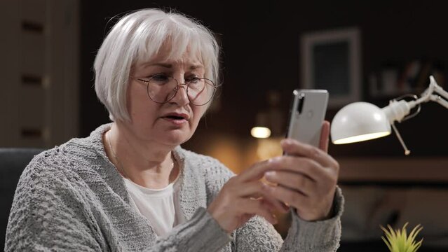 Sad senior woman with phone. Elderly gray-haired sad woman with glasses reads negative information on her phone, scrolls screen and nods sadly