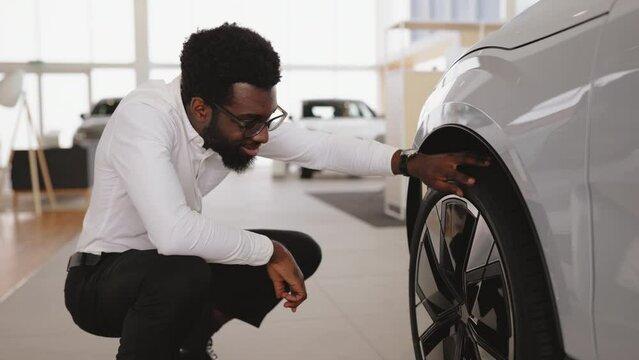 Smiling businessman wants to buy new automobile, while touch car tire wheel disk in showroom vehicle salon dealership store. Side view of customer, buyer, client in white shirt chooses auto.