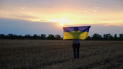 Happy lady walking on barley meadow and turning around with a raised over head flag of Ukraine. Ukrainian woman with a lifted blue-yellow banner on a beautiful sunset at background. End of war.