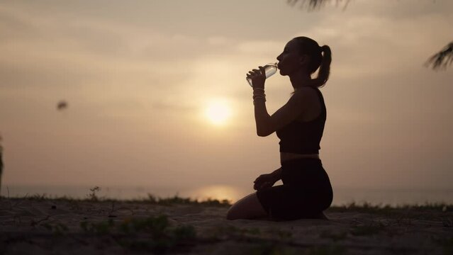 Woman drinking water after training. Fitness thirsty girl drinks water during workout pause, after a grueling run of a long sea ocean beach at sunset. Endurance sport, fitness, morning concept.