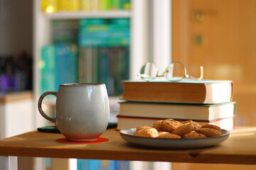 Cup of tea or coffee, pile of books, plate of cookies, reading glasses, e-reader and pen on the table. Colorful rainbow bookshelf in the background. Selective focus.