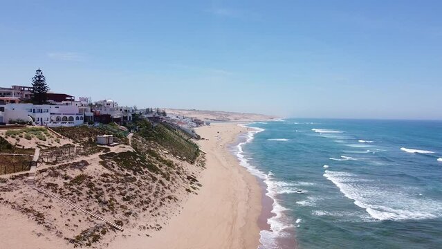 Pull-back drone footage of coastal white buildings on sand dunes on a sandy shore in Morocco