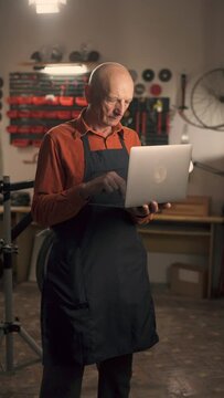 Serious repairman in bicycle shop with laptop computer in workshop. Elderly man Bike mechanic, professional mature technician in store or small business