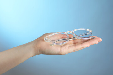 Woman holding glasses with transparent frame on light blue background, closeup