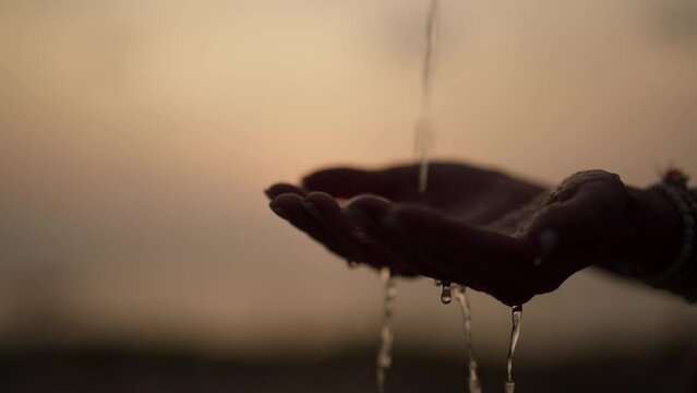 Girl puts palms under drops of water. Close-up woman's hands with drinking water pouring onto them on the beach against the backdrop of sea at sunset. Drought, refreshing moisture, ocean concept.