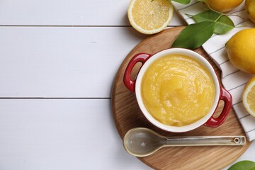 Delicious lemon curd in bowl, fresh citrus fruits and spoon on white wooden table, flat lay. Space...