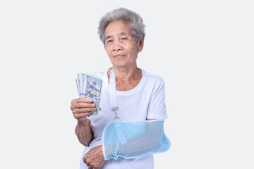A sen woman with broken arm and wearing a sling with a smiling face and holding dollars for insurance and paid care isolated on a white background. Old elderly female medical care. Health care concept © maemodnit13